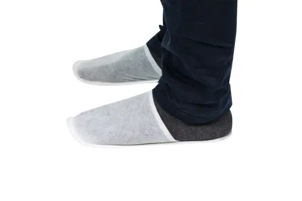 White disposable shoe covers worn indoors White disposable shoe covers protect dark footwear as a person stands against a plain white background