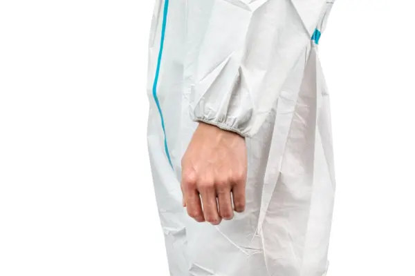 Close-up of a hand in a white protective suit A person’s relaxed hand hangs from a white protective coverall with an elastic cuff and blue seam against a plain background.