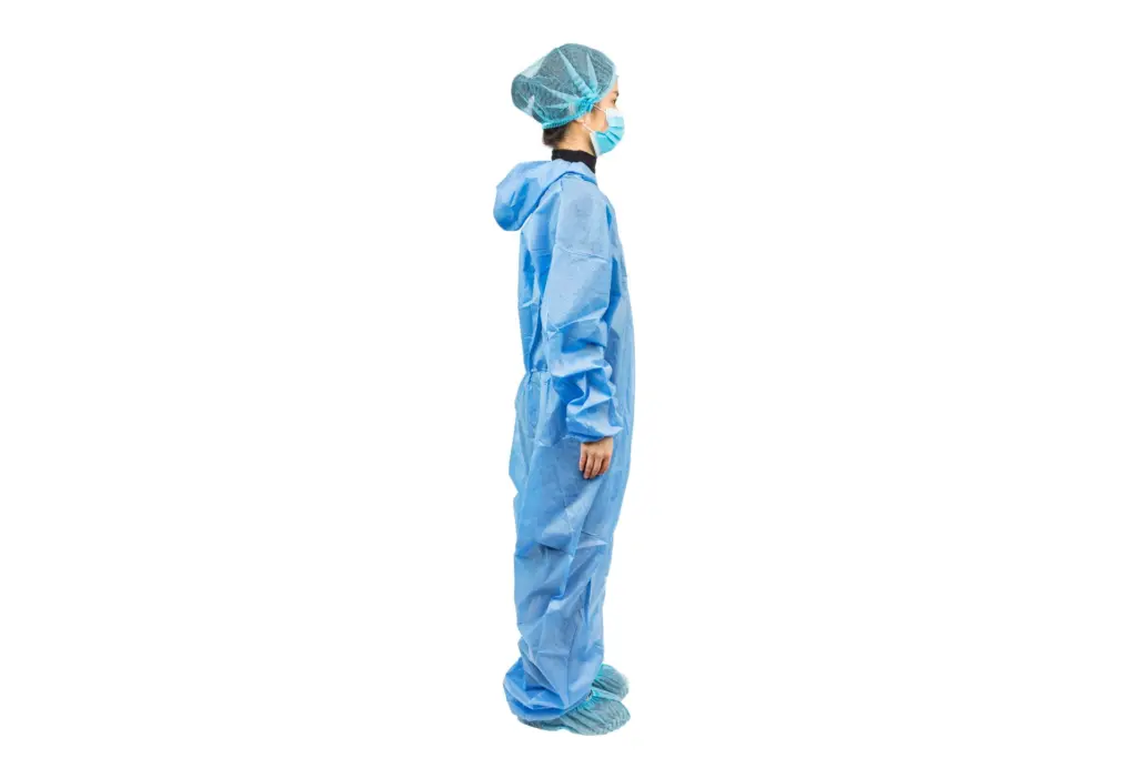Healthcare worker in blue protective coveralls, hair cover, face mask, and shoe covers stands in profile against white