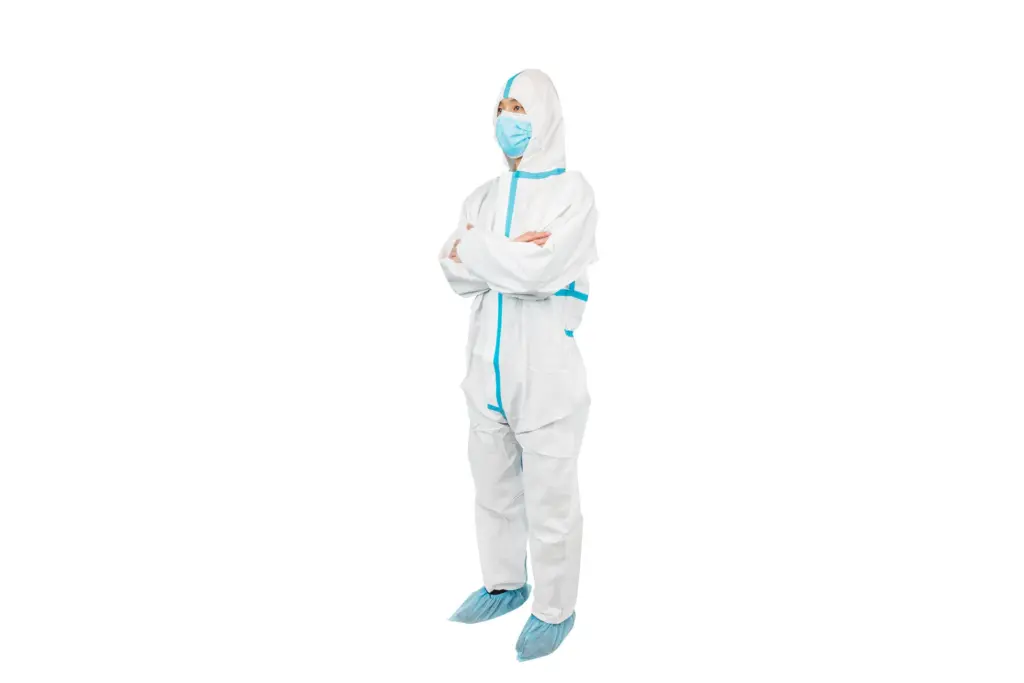 Person standing with folded arms in a white protective coverall, blue surgical mask, and blue shoe covers.