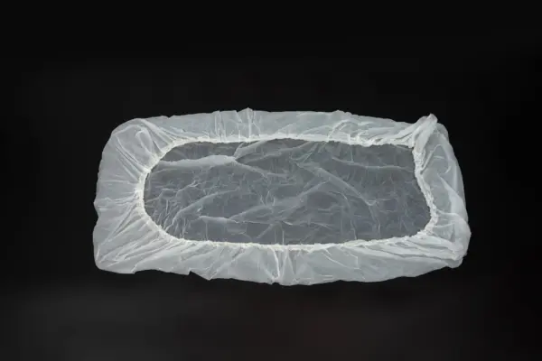 Transparent plastic cover with elastic edge Translucent rectangular plastic cover with gathered elastic edging laid flat against a black background.
