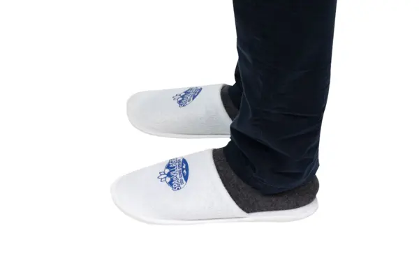 White house slippers worn with dark trousers White slip-on house slippers with dark trim worn on feet beneath dark trousers against a plain white background