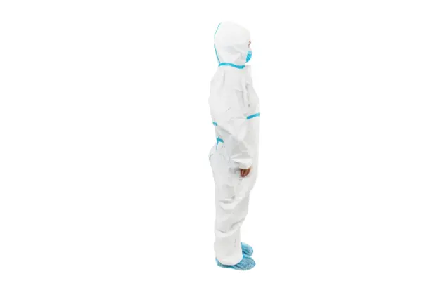 Protective coverall worn in side profile Person in a white protective coverall with blue mask and shoe covers stands in side profile against a plain background.