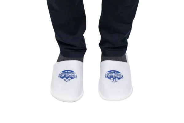Person wearing white closed-toe Spa Village slippers over dark socks and navy trousers against a plain white background.